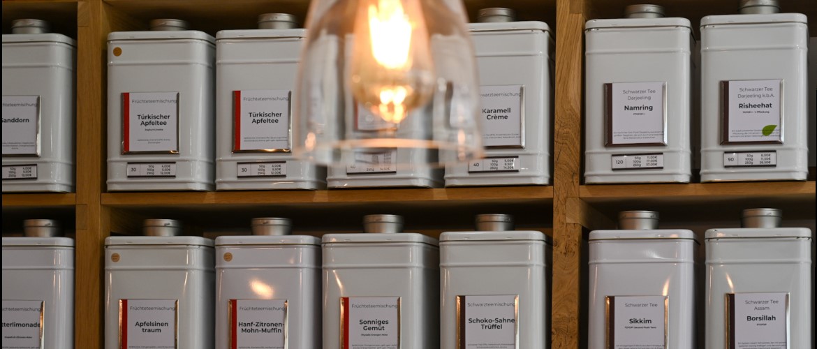 Zwei Regalböden mit ordentlich aufgereihten, weißen Teedosen, die im warmen Licht einer Lampe im Vordergrund stehen.