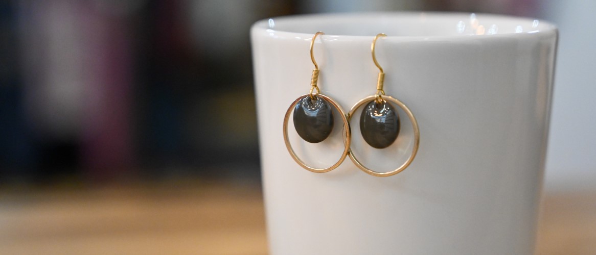 Ein Paar goldene Creolen-Ohrringe mit dunkelgrauen, glänzenden Emaille-Anhängern, die am Rand einer weißen Tasse hängen.