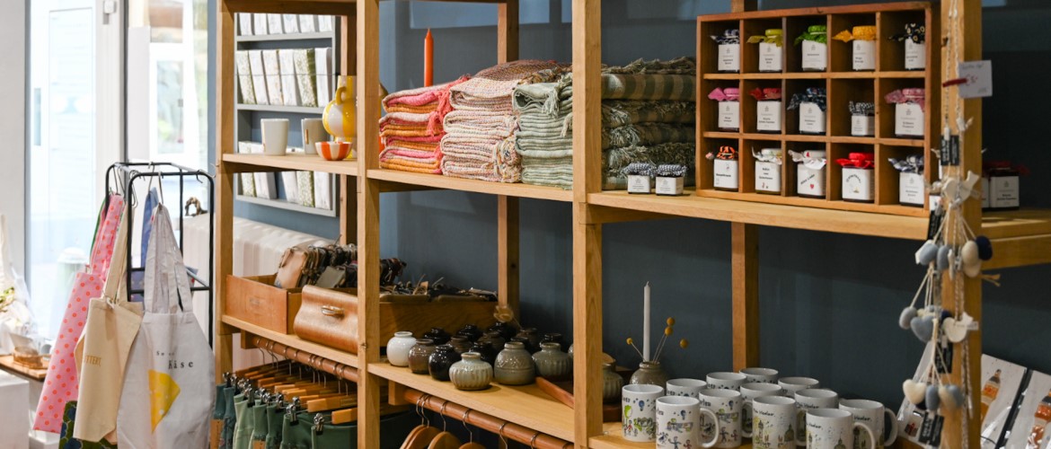 Ein großes Holzregal im Schauplatz Kevelaer, bestückt mit bunten Decken, kleinen Marmeladengläsern in einem Setzkasten, Tassen mit Stadtmotiven und verschiedenen Deko-Objekten.